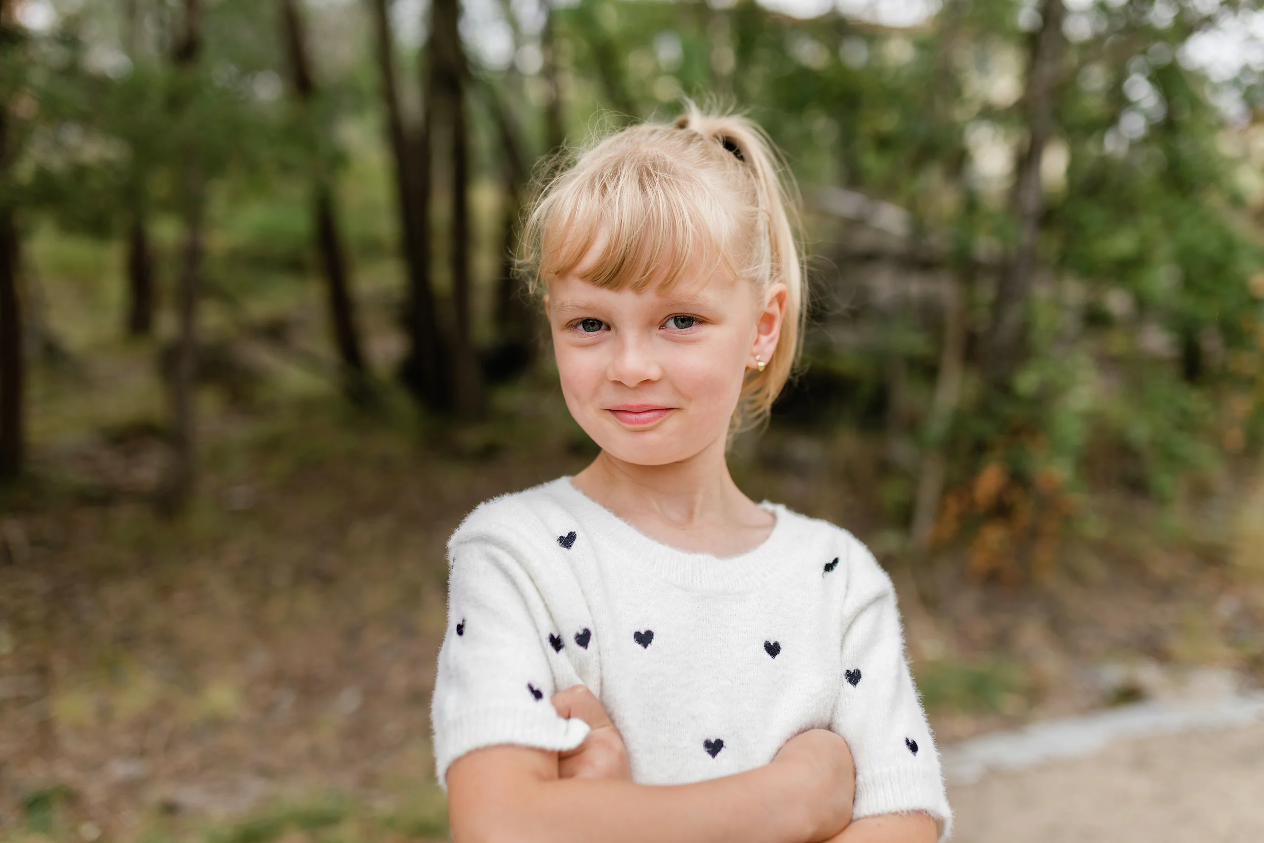 En ung blond pige med pandehår, armene over kors, iført en hvid hjertemønstret sweater, smilende udendørs i skovklædte omgivelser.
