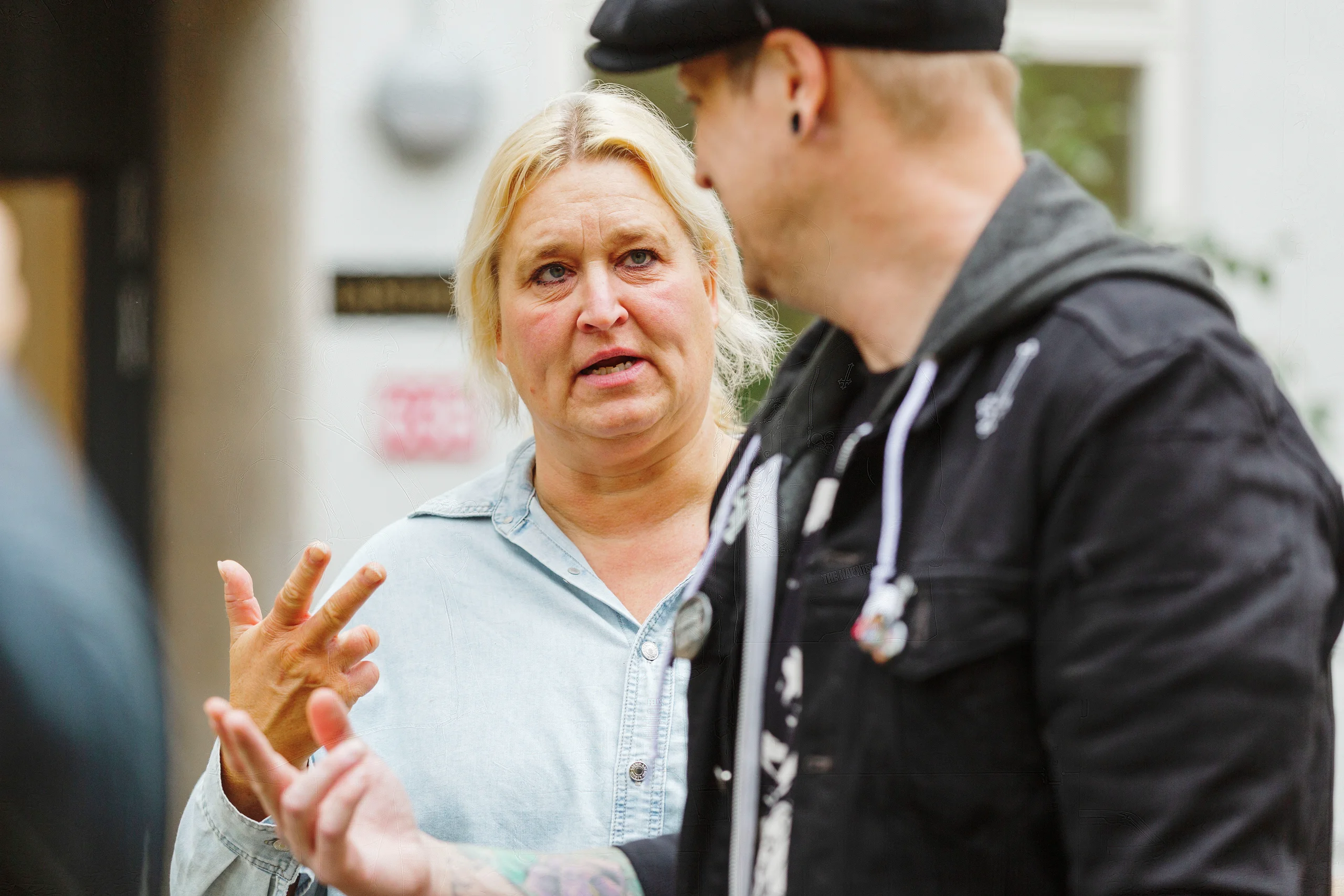 En blond kvinna i jeansskjorta gestikulerar medan hon pratar med en person i svart jacka utomhus.