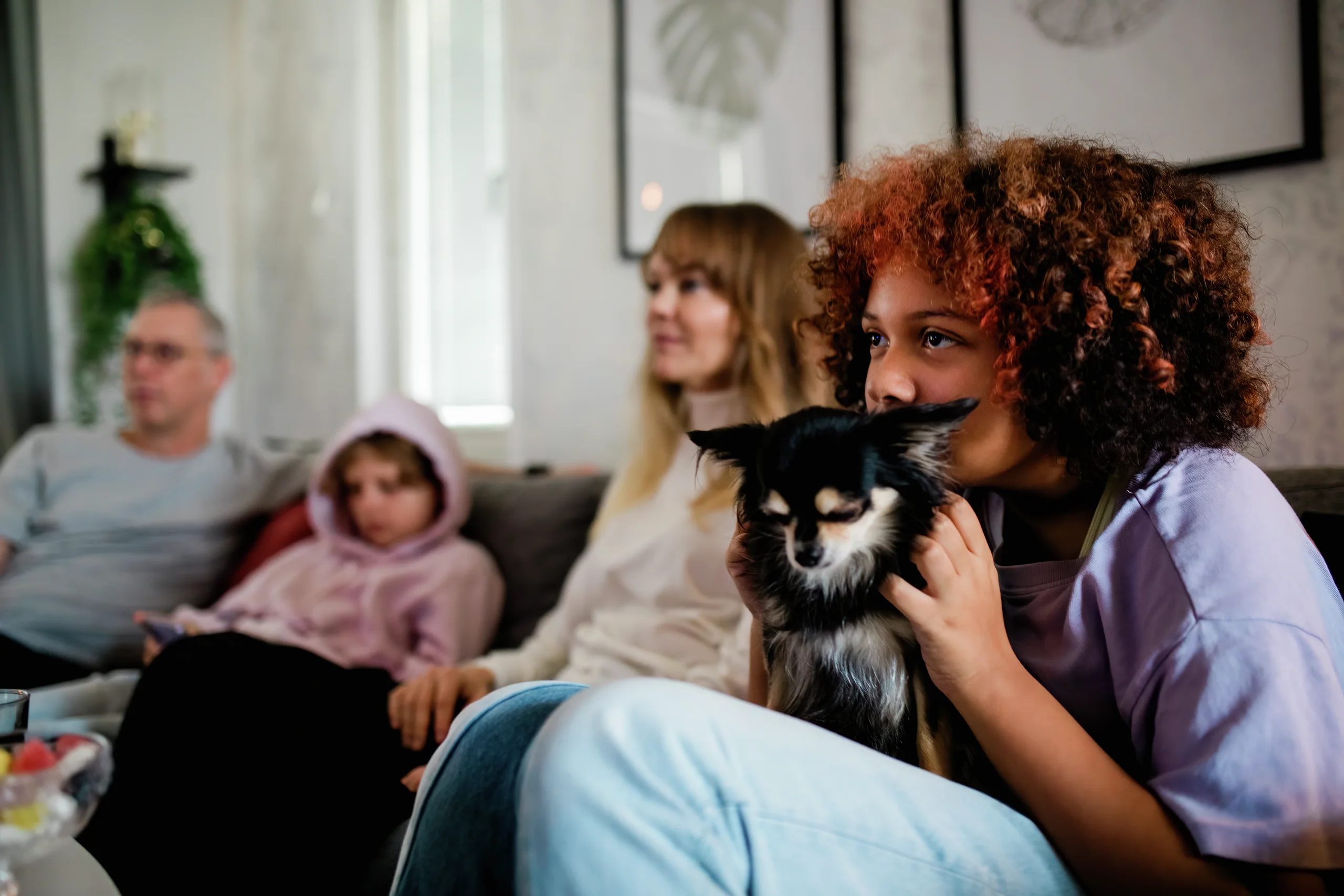 En lockig rödhårig flicka håller en liten svart hund medan familjemedlemmar sitter tillsammans i en soffa.