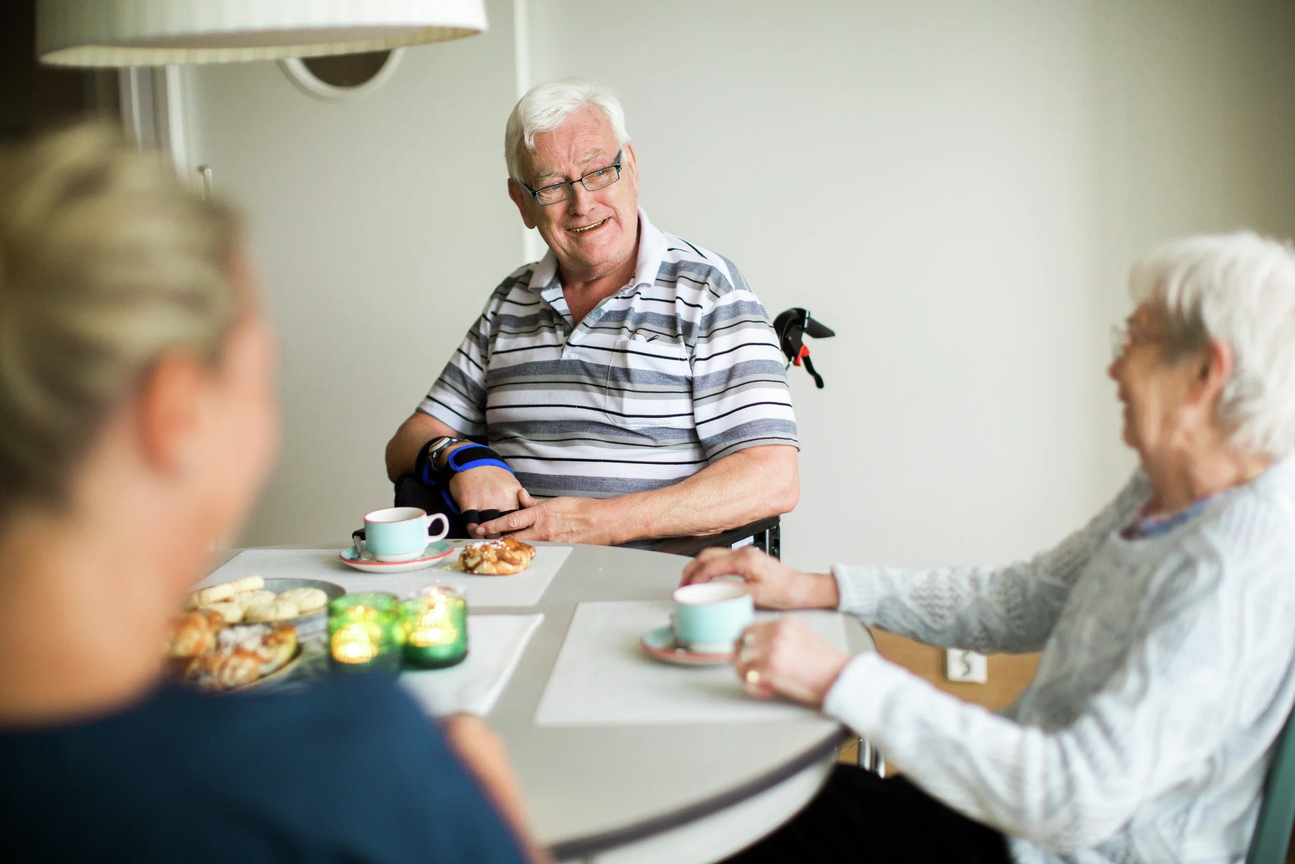 Tre äldre personer sitter runt ett bord och delar kaffe och bakverk, en man bär blodtrycksmätare.